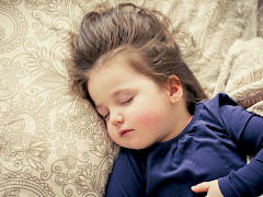 Lapsi nukkuu beigen tyynyn päällä. Lapsella on ruskeat hiukset ja tummansininen paita. Käsi on vatsan päällä ja korvassa on pieni korvakoru.