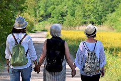 Kolme naista selin kesäisessä maisemassa kävelemässä. Kaikilla on reput selässä. Kahdella reunimmaisella naisella on hattu päässä.