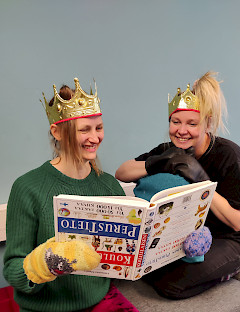 Kuvassa kaksi naista lukevat väärinpäin olevaa kirjaa ja nauravat. Heillä on kruunut päässä ja hanskat kädessä.