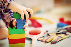 Lapsi rakentaa värikkäill dublo-palikoilla tornia. Vain lapsen kädet näkyvät kuvassa ja lattialla muita leluja.