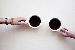 Kaksi kättä, jotka pitävät valkoisia kahvikuppeja, joissa kahvia, vastakkain. Toisella on ranteessa koruja.