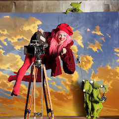 Kuvassa kolme hahmoa ja kamera. Yksi nainen, jolla punaista päällä ja hattu. Nainen kiivennyt jalalliseen kameraan. Vieressä on kaksi vihreeä pehmohahmoa. Kuvaaja Marko Mäkinen.