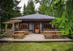 Hirsimökki, jossa terassi edessä ja pöytiä. Ympärillä puita.